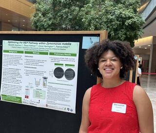 A woman with curly brown hair stands in front of a science presentation poster, with a tree in an open room behind her