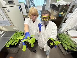 Woman with blonde hair grins and holds a plant out to the camera beside another person in a lab coat and goggles