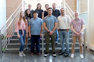 Ten People stand in front of stairs and a bright window