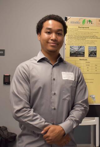 A man with short curly black hair stands in front of a science presentation poster