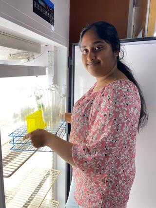 A woman with long black hair stands in front of an open fridge holding a tray full of empty tubes.