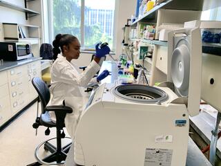 A woman with black hair in a lab coat sits in a lab, holding a pipette and a plastic tube