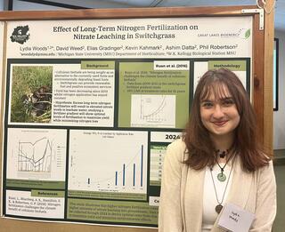 A woman with brown hair and bangs stands in front of a scientific presentation poster