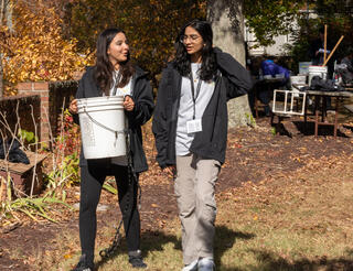 Two girls walk outdoors on an autumn day, surrounded by foliage and fallen leaves. The girl on the left is carrying a 5-gallon plastic bucket. Both are smiling and appear to be in conversation. Garden tools and other supplies are visible in the background.