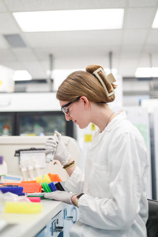 Peterson wears a white lab coat and gloves as she uses a pipette in a lab