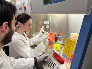 White using a pipette in the fume hood as a man in glasses and PPE looks on