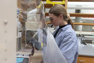 Woman in blue and white lab coat faces a clear enclosure with rubberized gloves built in