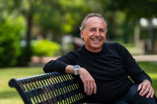 Man in a black shirt sitting on an outdoor bench surrounded by greenery. One arm is on the back of the bench and he is turned to face the camera.