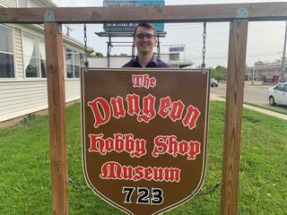 A man stands behind a wooden sign reading "The Dungeon Hobby Shop Museum" in red letters.
