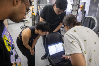 Four people in a laboratory lean in together to view data on a laptop screen. Behind them stands a large, white automated workstation with circular portals and black gloves.