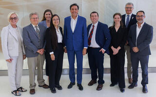 Nine people in business attire pose smiling for a group photo in a bright hallway, with a tall man in a bright blue suit standing at the center.