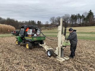 A man stands in a barren farm field with a large mechanical drill attached to a four-wheeler