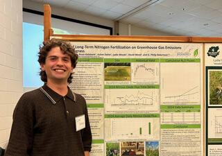 A man with short curly brown hair stands in front of a science presentation poster