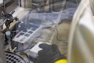 Closeup of a hand in a black rubber glove holding a glass container and a pipette