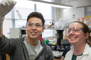 Two smiling researchers wearing safety glasses examine a small test tube held up to the light in a chemistry laboratory.
