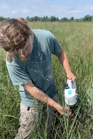 A young student holds a machine that measures photosynthesis up to a blade of grass