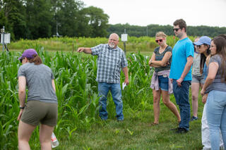 A group of people stand next to a plot of waist-high corn looking as a man gestures toward the field