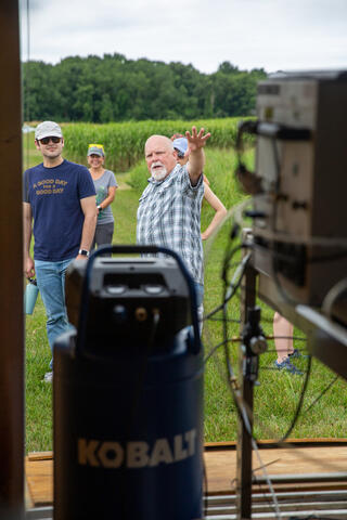 Man in blue checkered shirt standing in field gestures towards equipment in an enclosure as seen from inside the enclosure