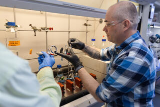 Man in blue checkered shirt and black rubber gloves holds a piece of lab equipment under an exhaust hood