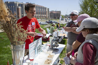 A researcher in a red "Explore Science" t-shirt explains a biofuel production display — featuring a microscope, yeast samples, and informational posters — to visitors at an outdoor public science outreach event on a university campus.