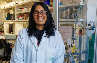A smiling researcher in a white lab coat and glasses stands in front of a fume hood and chemical shelving in a chemistry laboratory.