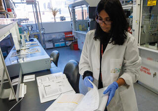 A researcher in a lab coat and blue gloves reviews a lab notebook at a benchtop, with analytical instruments and a fume hood visible in the background.