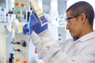 A researcher in a white lab coat, safety glasses, and blue gloves pipettes a sample into a microcentrifuge tube in a well-stocked microbiology laboratory.