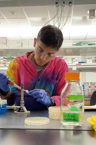 Salvador Castillo, a young man in a tye-dye lab coat, pipettes at a lab bench