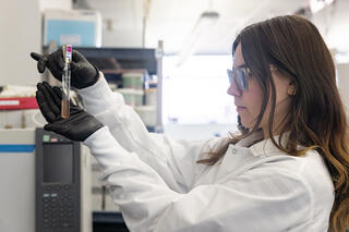 A woman in a white lab coat and black latex gloves holds up a test tube filled with amber liquid