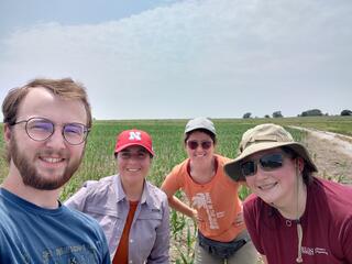 Four smiling researchers pose for a selfie in a young cornfield on an overcast day, one wearing a University of Nebraska cap and another an Elon University shirt.