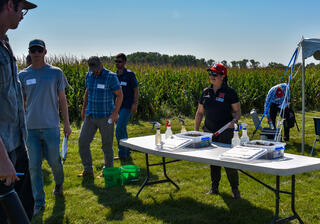 A University of Nebraska extension educator in a red "N" cap leads an outdoor field demonstration for a small group of attendees beside a cornfield, with soil samples and spray bottles displayed on a folding table.