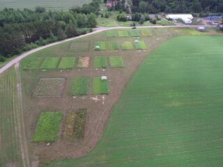 The image shows an aerial view of a rural farming area. There is a dirt road running across the top of the image. Several small plots of land are visible, some with planted crops of varying shades of green and yellow.