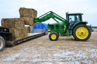 A green tractor loads large bales of brown material onto a trailer