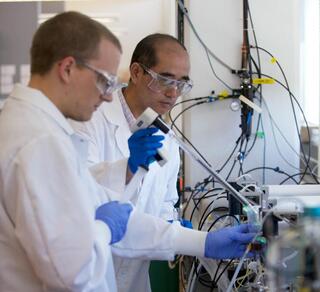 Two men in white lab coats and blue rubber gloves inject liquid into a machine